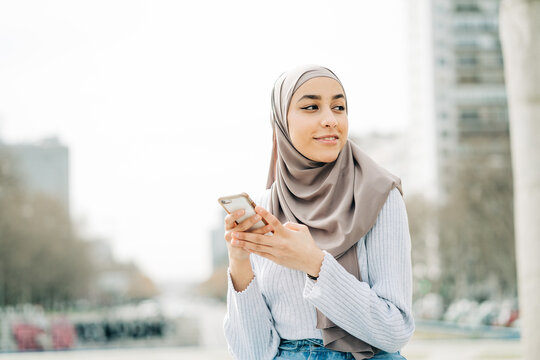 Young ethnic female in hijab standing in city and messaging on mobile phone