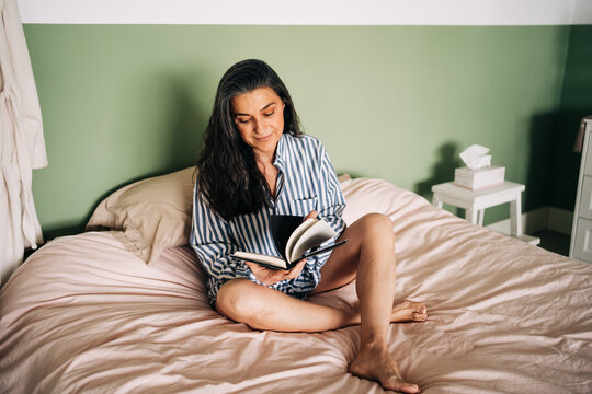 Relaxed mature Hispanic woman in casual clothes sitting in bed and reading interesting book