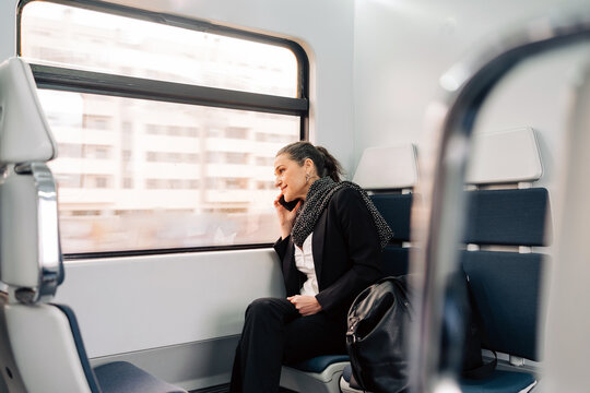 Side view of content female sitting on passenger seat and looking out window while having phone call in modern train