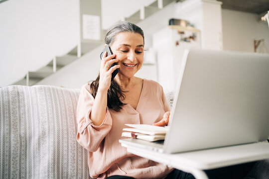 Concentrated middle aged female having phone call and taking notes in notepad while looking at screen of laptop in light room at home