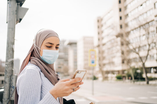 Side view of ethnic female wearing headscarf and protective mask standing on street in city on sunny day and looking at screen of her mobile
