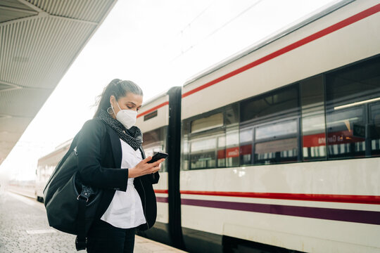 Serious female with bag wearing protective respirator text messaging on cellphone while standing on railway station near modern train during trip