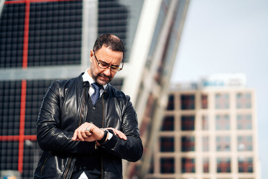 Serious mature Hispanic male executive in formal apparel and eyewear checking time on wristwatch in town