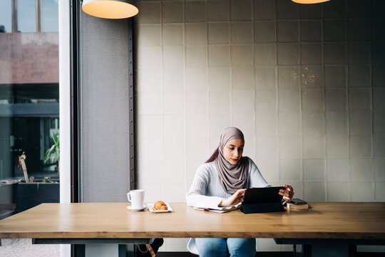 Content Muslim female in hijab and talking on video chat via tablet while sitting at table in cafe