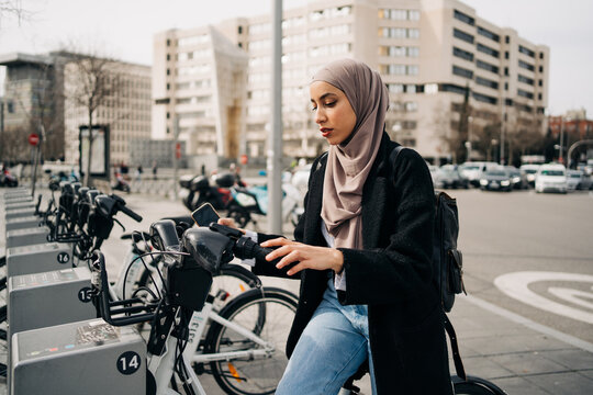 Side view of Muslim female in headscarf using bicycle sharing system in city