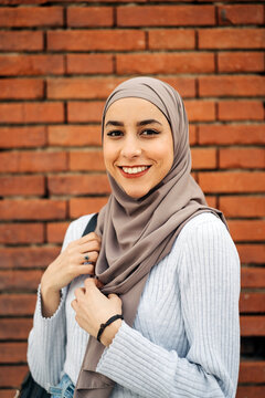 Charming Muslim female in traditional headscarf and trendy clothes standing on street against brick wall and looking at camera