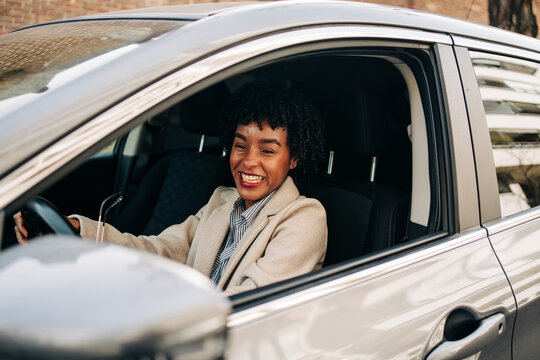 Side view of cheerful African American female driver in fashionable outfit smiling while driving modern automobile on the street