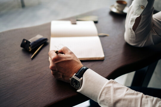 Crop anonymous businessman in wristwatch with pen and open journal working at cafeteria table in daylight