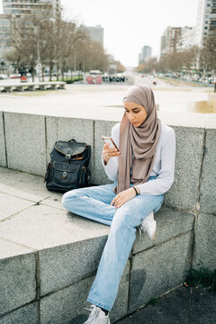 Young ethnic female in hijab sitting on stone border in city and messaging on mobile phone