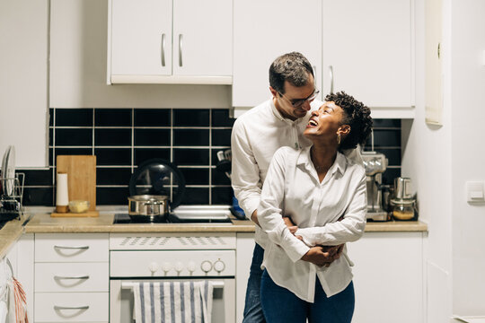 Delighted Man Embracing Smiling Black Woman From Behind While Standing In Kitchen And Looking At Each Other