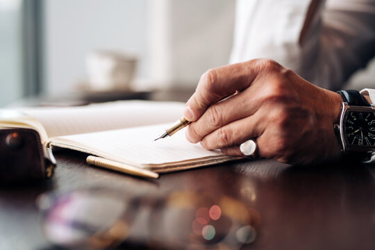 Crop anonymous businessman in wristwatch with pen and open journal working at cafeteria table in daylight