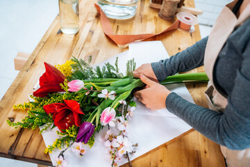 From above cropped unrecognizable female florist in casual clothes and apron arranging elegant bouquet of assorted flowers in store