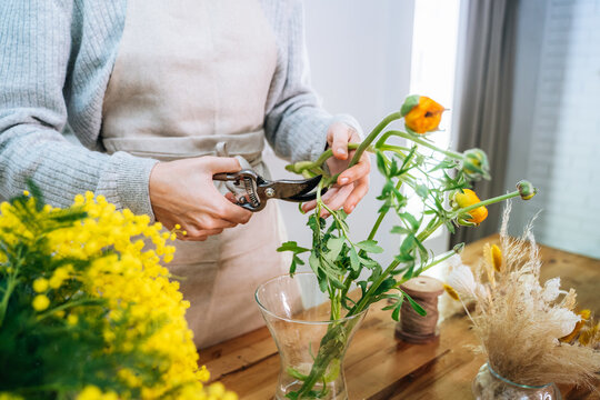 Crop unrecognizable young female florist in casual clothes cutting stems of fresh bright flowers with pruning snips while arranging bouquet in glass vase