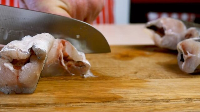 Cook cuts carcass of hake or pollock fish into pieces with sharp knife to prepare delicious food, on wooden cutting board in domestic kitchen. Recipe of fried fish. Selective focus.