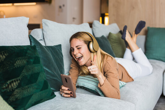 Joyful female lying on sofa and enjoying music in headphones while winking and pointing at camera