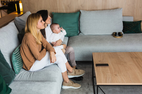 Side view high angle of content couple of lesbian women sitting on sofa with cute baby and watching movie together in living room