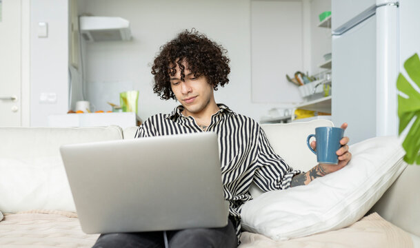 Young hipster male with curly hair browsing internet on netbook while resting on couch in house room