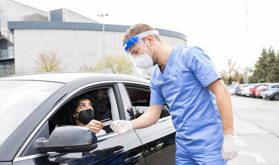 Male doctor in protective uniform, latex gloves and face shield checking African American female patient Identification card before vaccine procedure inside the car on a drive through mobile clinic during coronavirus outbreak