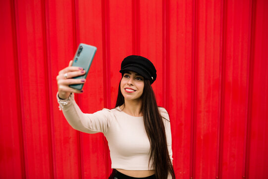 Cheerful trendy female entrepreneur standing against red wall in city and taking self shot on smartphone