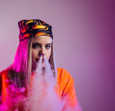 Cool female in street style outfit smoking e cigarette and exhaling smoke through nose on purple background in studio with pink neon illumination