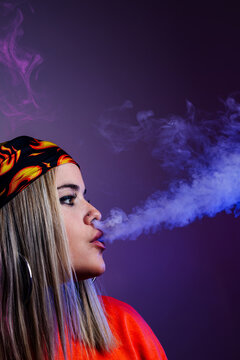 Side view of cool female in street style outfit smoking e cigarette and exhaling smoke through nose on purple background in studio with pink neon illumination