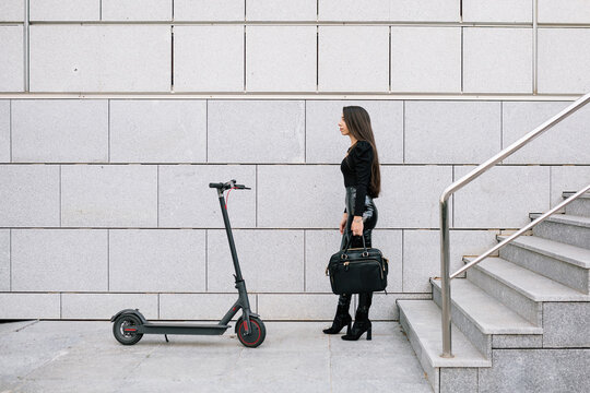 Side view of confident trendy female entrepreneur standing near contemporary electric scooter on city street