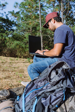 From below nomad sitting on stone near belongings and using netbook while working remotely