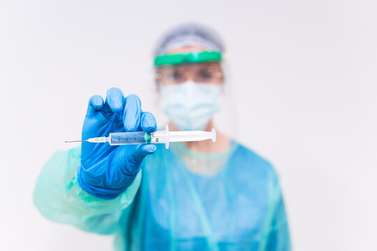 Blurred unrecognizable medic in protective face shield mask and latex gloves with vial of coronavirus vaccine and syringe showing to camera while standing in hospital room