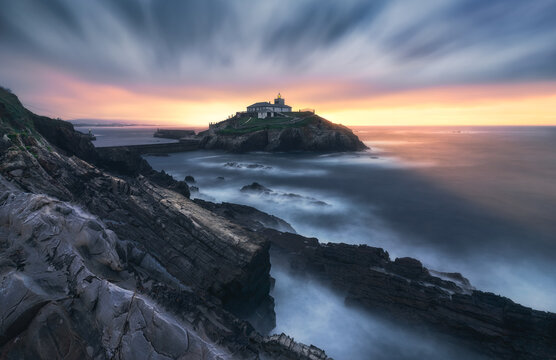 Breathtaking scenery of rocky island with lighthouse located in ocean near rocky coast in Faro Tapia de Casariego in Asturias in Spain under cloudy sky at sunrise