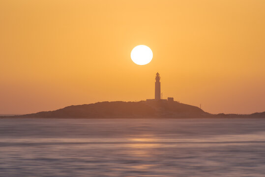 Picturesque scenery of rippling water of ocean washing coast with high rise lighthouse placed in Faro de Trafalgar in Cadiz in Spain under bright orange sky at sunrise