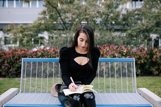Stylish female entrepreneur sitting on bench and taking notes in organizer while working in urban park