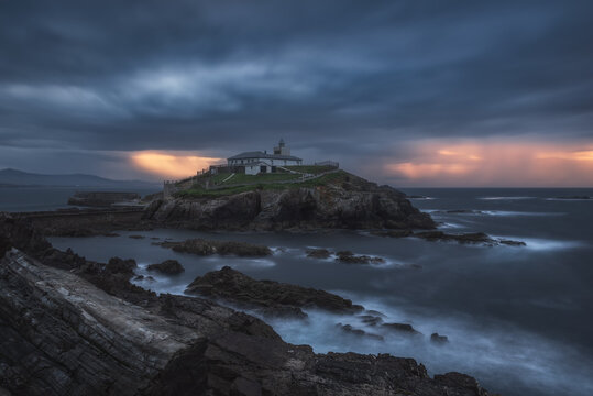 Breathtaking scenery of rocky island with lighthouse located in ocean near rocky coast in Faro Tapia de Casariego in Asturias in Spain under cloudy sky at sunrise