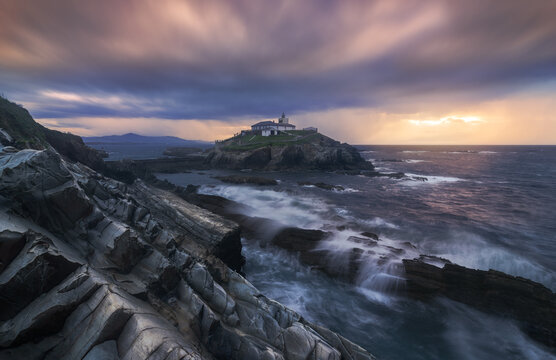 Breathtaking scenery of rocky island with lighthouse located in ocean near rocky coast in Faro Tapia de Casariego in Asturias in Spain under cloudy sky at sunrise