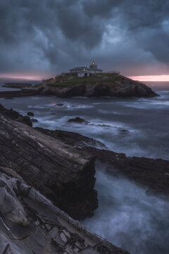 Breathtaking scenery of rocky island with lighthouse located in ocean near rocky coast in Faro Tapia de Casariego in Asturias in Spain under cloudy sky at sunrise