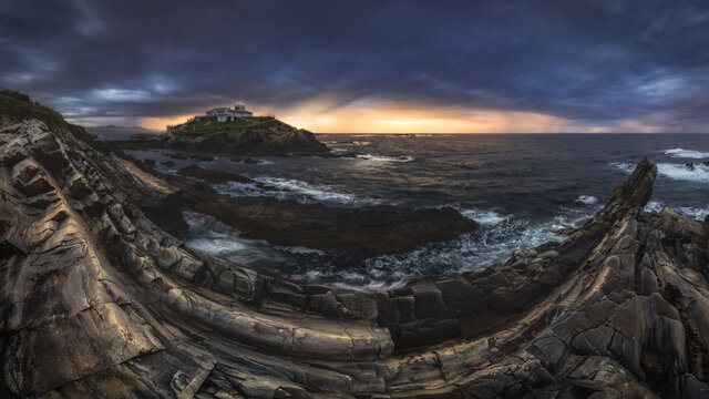 Breathtaking scenery of rocky island with lighthouse located in ocean near rocky coast in Faro Tapia de Casariego in Asturias in Spain under cloudy sky at sunrise