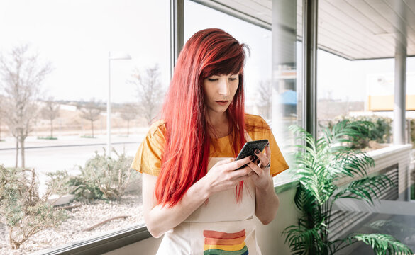 Young homosexual female with dyed hair in apron text messaging on cellphone in light house