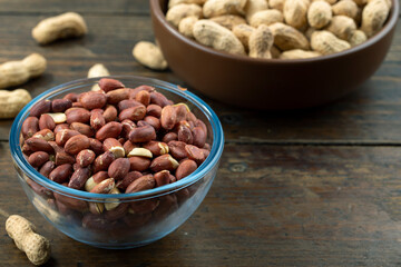 Peanuts in their husks in a glass bowl and peanuts in their skins are scattered on the table. Space for text.