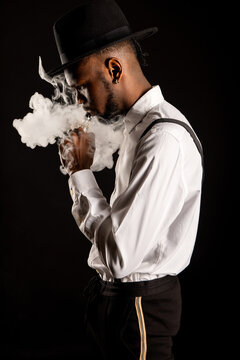 Side view of masculine African American male in white shirt and hat exhaling vapor while smoking e cigarette