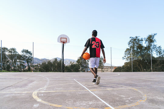 Back view of unrecognizable African American male streetball player in uniform with ball on basketball court