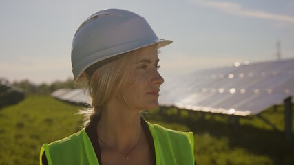 Portrait of happy female engineer in protective helmet while looking to camera. Handsome woman in uniform smiling while standing at solar power farm. Concept of green energy