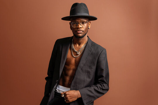 Young masculine unshaven African American male with naked abdomen in jacket looking at camera on brown background