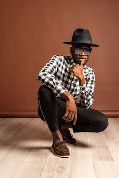 Young cool African American male model in checkered shirt and hat looking at camera while crouched on brown background