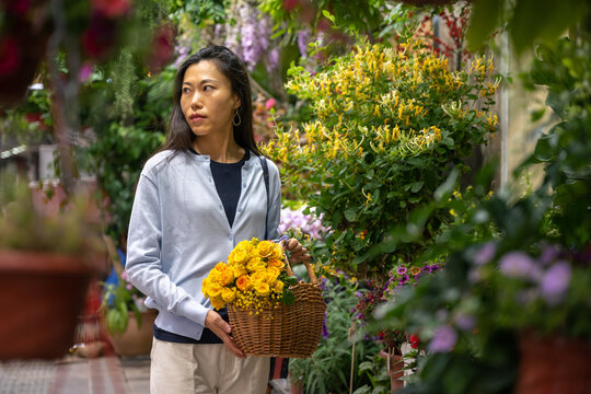 Beautiful Asian girl buying flowers in flower shop while carrying a wicker basket with yellow flowers.