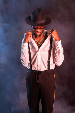 Young masculine ethnic male model in hat and trousers standing while looking at camera on black background with smoke