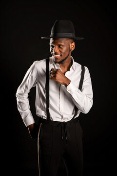Young masculine ethnic male model in hat and trousers standing while looking away on black background with smoke