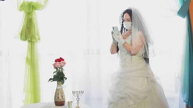 A Jewish bride in a wedding dress and a veil wearing a medical mask in a synagogue hall answers an SMS before the chuppah ceremony