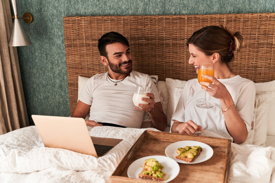 Happy young multiethnic couple in casual clothes having breakfast and watching movie on laptop while sitting in comfortable bed during lazy weekend at home - Powered by Adobe