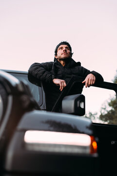 From below adventurer leaning on the door of an off-road car while looking away before starting the journey