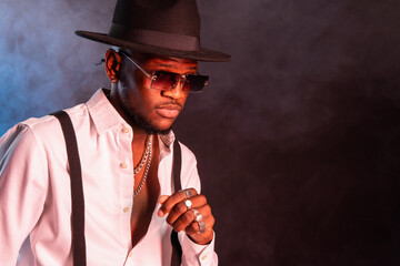 Young masculine ethnic male model in hat and trousers standing while looking away on black background with smoke