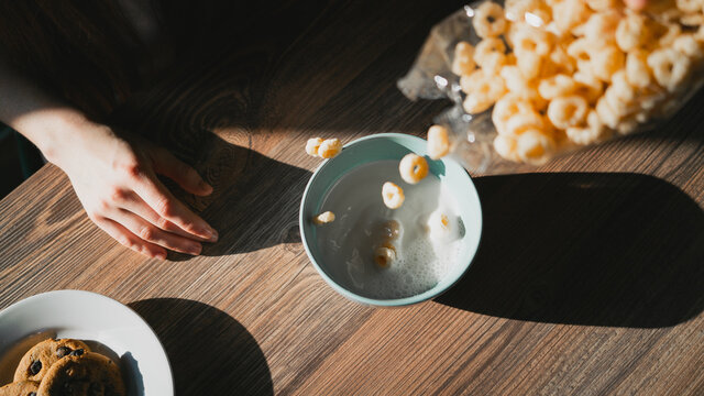 Crop unrecognizable person pouring milk into bowl with delicious corn rings for breakfast on wooden table